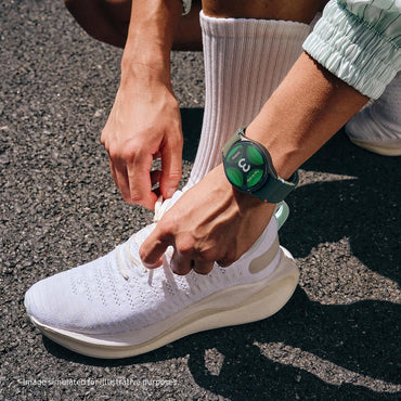 Person tying shoelaces on a white sneaker with a smartwatch on the wrist, standing on a pavement.